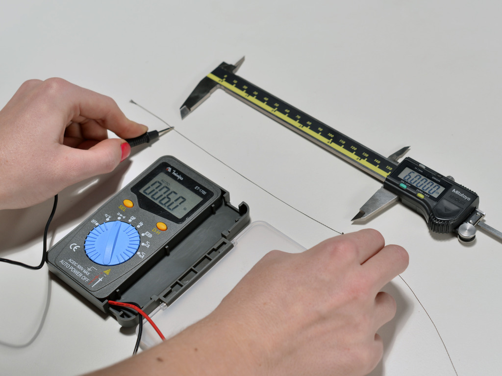 Image showing a pair of hands holding probes touching a length of conductive thread. The probes are attached to a multimeter, which reads the resistance of the thread.